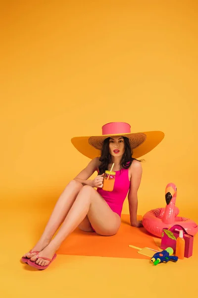 Красива жінка в солом'яному капелюсі і купальному костюмі тримає скло з апельсиновим соком біля надувного кільця і водяної гармати на жовтому — стокове фото