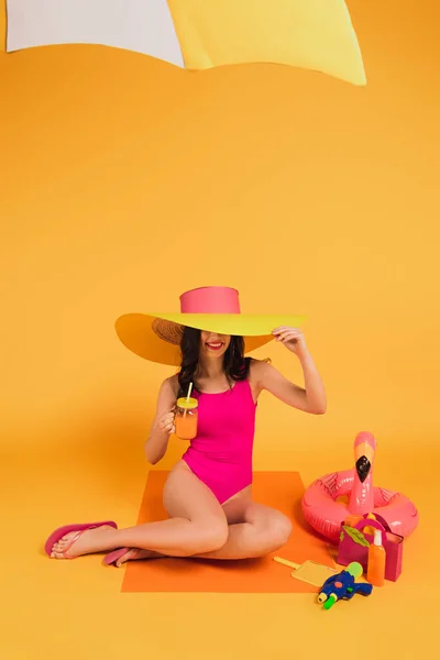 Mujer feliz en sombrero de paja y traje de baño sosteniendo vidrio con jugo de naranja cerca de anillo inflable y pistola de agua en amarillo - foto de stock