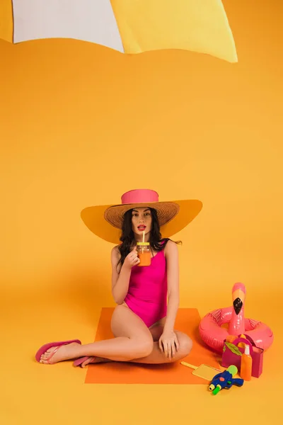 Atractiva chica en sombrero de paja y traje de baño celebración de vidrio con jugo de naranja cerca de anillo inflable y pistola de agua en amarillo - foto de stock