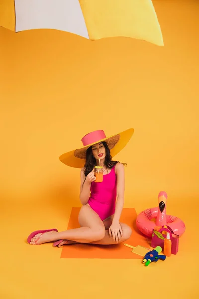 Menina atraente em chapéu de palha e maiô segurando vidro com suco de laranja perto da pistola de água e anel inflável no amarelo — Fotografia de Stock