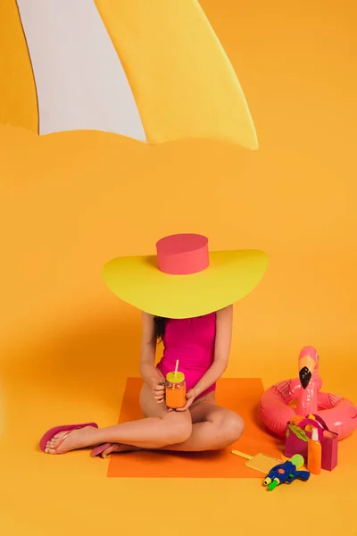 Niña en sombrero de paja y traje de baño rosa celebración de vidrio con jugo de naranja cerca de la pistola de agua y anillo inflable en amarillo - foto de stock