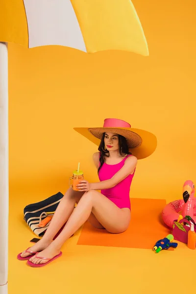 Elegante ragazza in cappello di paglia e costume da bagno in possesso di vetro con succo d'arancia vicino pistola ad acqua e anello gonfiabile su giallo — Foto stock