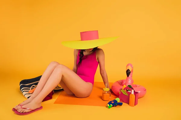 Stylish woman in straw hat and pink swimsuit sitting near glass with orange juice, water gun and inflatable ring on yellow — Stock Photo