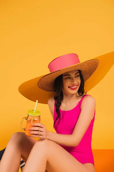Весела дівчина в солом'яному капелюсі і купальнику тримає скло з апельсиновим соком на жовтому — стокове фото