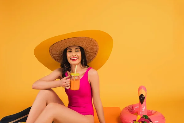 Fröhliches Mädchen mit Strohhut und Badeanzug, das Glas mit Orangensaft in der Nähe von rosa Schwimmring auf gelb hält — Stockfoto