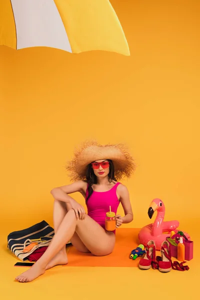 Elegante donna in cappello di paglia, occhiali da sole e costume da bagno rosa in possesso di vetro con succo d'arancia vicino pistola ad acqua e anello gonfiabile su giallo — Foto stock