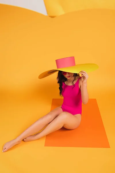 Selective focus of stylish woman in straw hat and swimsuit sitting on yellow — Stock Photo