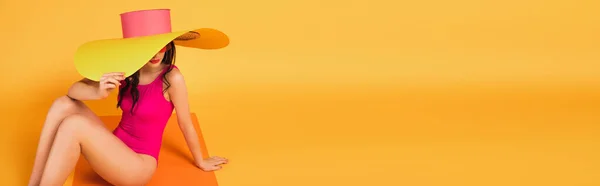 Orientación panorámica de la mujer con estilo en sombrero de paja y traje de baño que cubre la cara mientras está sentado en amarillo - foto de stock