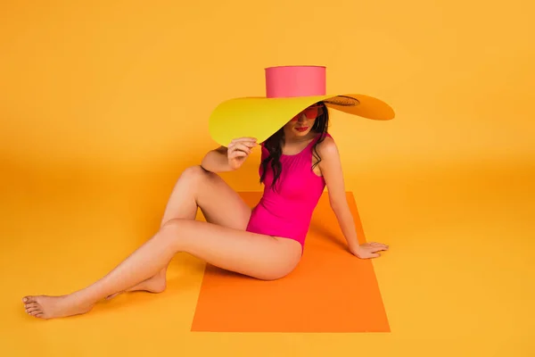 Chica con estilo en sombrero de paja y traje de baño que cubre la cara mientras está sentado en amarillo - foto de stock