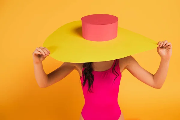 Fille élégante en maillot de bain rose couvrant visage avec chapeau de paille sur jaune — Photo de stock