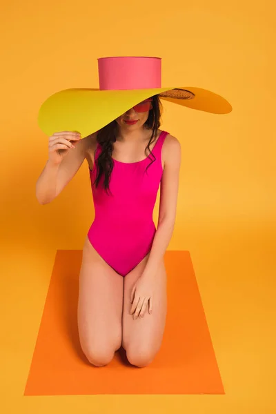 Stylish woman in pink bathing suit covering face with straw hat on yellow — Stock Photo