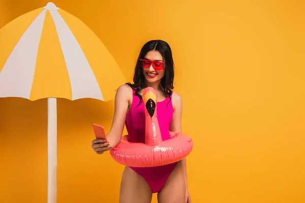 Happy girl in sunglasses and swimsuit standing with inflatable ring and taking selfie on yellow — Stock Photo
