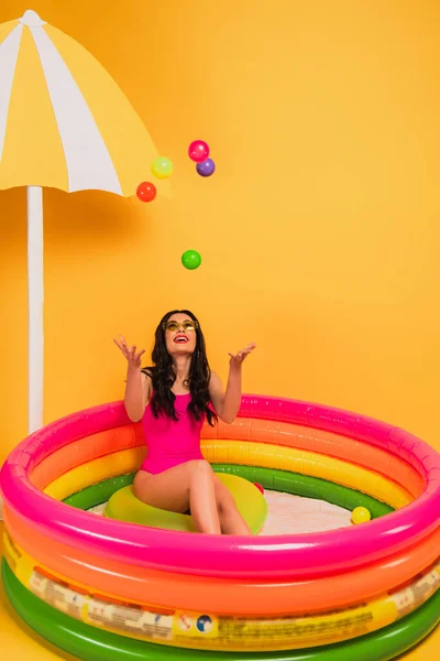 Menina feliz jogando bolas de ar enquanto sentado na piscina inflável no amarelo — Fotografia de Stock