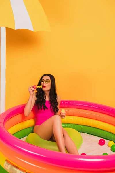 Young woman in swimsuit blowing soap bubbles while sitting in inflatable pool on yellow — Stock Photo
