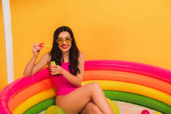 Glückliche Frau im Badeanzug hält Flasche mit Seifenblasen, während sie im aufblasbaren Pool auf gelb sitzt — Stockfoto