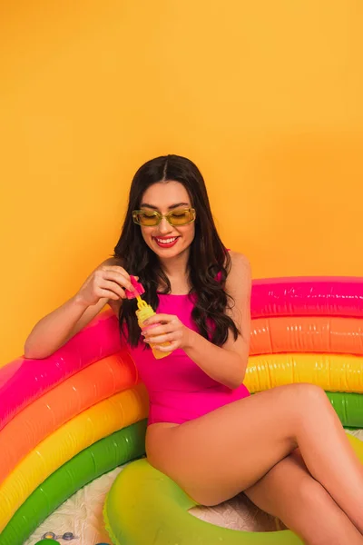 Fröhliche Frau im Badeanzug hält Flasche mit Seifenblasen, während sie im aufblasbaren Pool auf gelb sitzt — Stockfoto