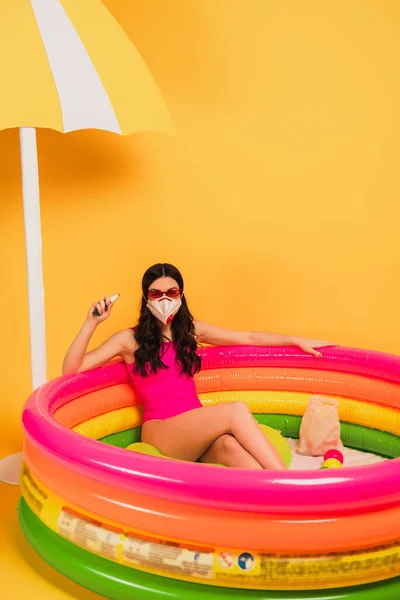 Femme en maillot de bain, lunettes de soleil et masque médical assis dans la piscine gonflable et tenant désinfectant sur jaune — Photo de stock