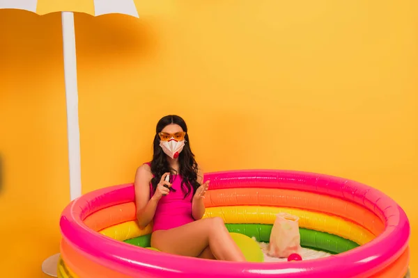 Молода жінка в купальнику, сонцезахисних окулярах і медичній масці сидить в надувному басейні і тримає дезінфікуючий засіб на жовтому — стокове фото
