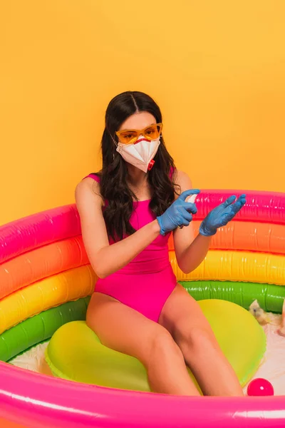 Молода жінка в купальнику, сонцезахисних окулярах, латексних рукавичках і медичній масці, сидить в надувному басейні і застосовує дезінфікуючий засіб на жовтому — стокове фото