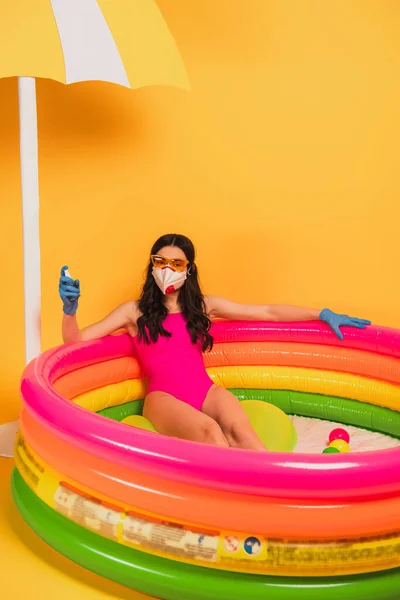 Молодая женщина в купальнике, солнцезащитных очках, латексных перчатках и медицинской маске сидит в надувном бассейне и держит дезинфицирующее средство на желтом — стоковое фото