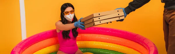 Horizontal crop of delivery man giving pizza boxes to woman in swimsuit, medical mask and sunglasses on yellow — Stock Photo