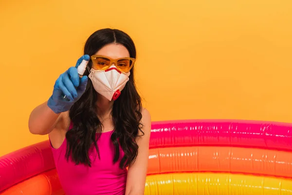 Jovem mulher em maiô, óculos de sol e máscara médica sentado na piscina inflável e segurando desinfetante no amarelo — Fotografia de Stock
