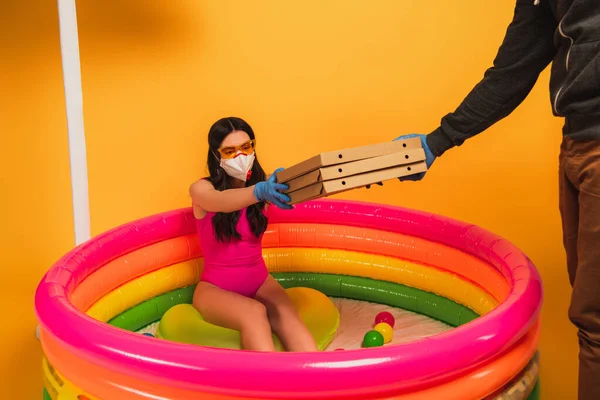 Delivery man giving pizza boxes to woman in swimsuit, medical mask and sunglasses on yellow — Stock Photo