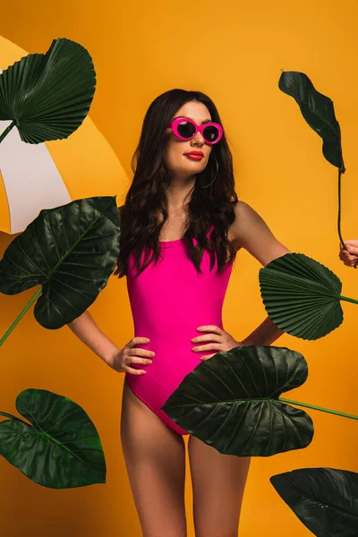 Ragazza elegante in occhiali da sole in piedi con le mani sui fianchi vicino foglie di palma verde su giallo — Foto stock