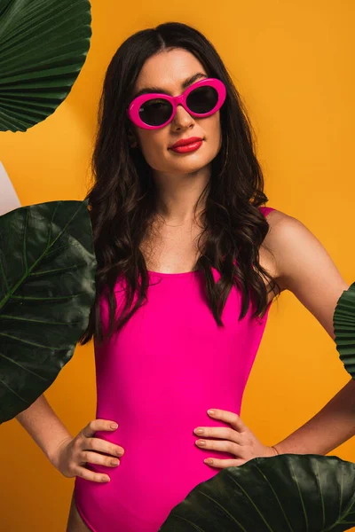 Stylish girl in sunglasses and swimsuit standing with hands on hips near green palm leaves on yellow — Stock Photo