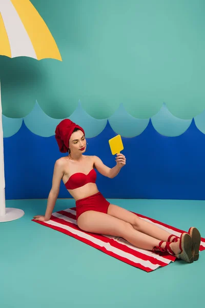 Mujer alegre en toalla roja y traje de baño mirando el espejo cerca de las ondas de corte de papel azul - foto de stock