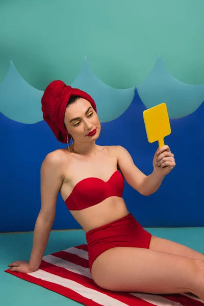 Hermosa mujer en toalla roja y traje de baño mirando el espejo cerca de las ondas de corte de papel azul - foto de stock