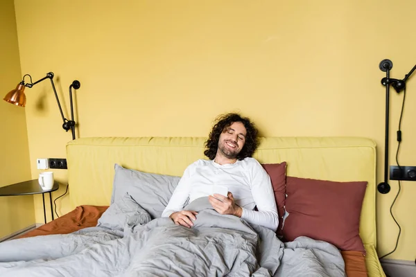 Happy young man smiling while chatting on smartphone in bed — Stock Photo