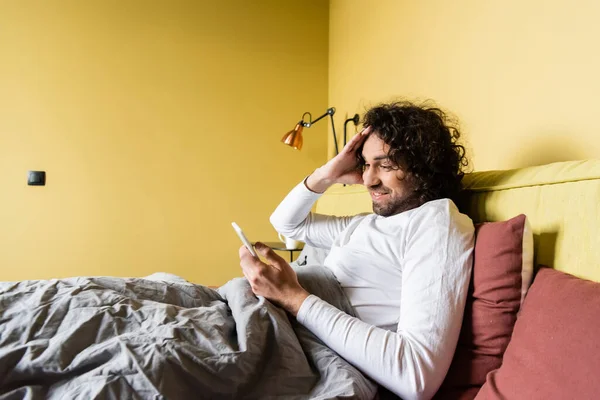 Шокований молодий чоловік торкається волосся під час спілкування на смартфоні в ліжку — стокове фото