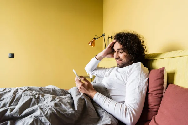 Conmocionado joven tocando el pelo mientras charlaba en el teléfono inteligente en la cama - foto de stock