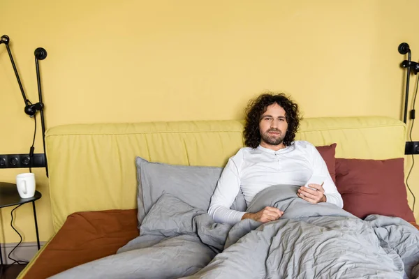 Hombre serio, rizado mirando a la cámara mientras chatea en el teléfono inteligente en la cama - foto de stock