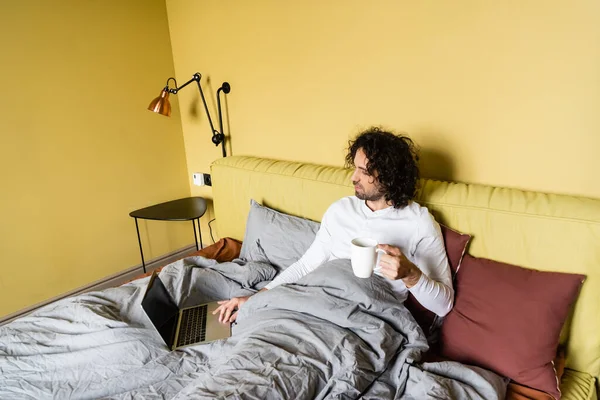 Vista de ángulo alto del freelancer rizado utilizando el ordenador portátil con pantalla en blanco mientras sostiene la taza de café en la cama - foto de stock