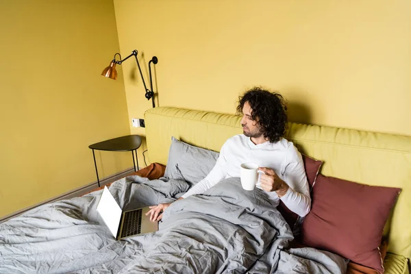 Vista de ángulo alto del joven freelancer utilizando el ordenador portátil con pantalla blanca mientras sostiene la taza de café en la cama - foto de stock