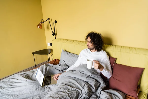 KYIV, UCRANIA - 25 DE ABRIL DE 2020: vista de alto ángulo del joven freelancer usando el ordenador portátil con el sitio web de Facebook mientras sostiene la taza de café en la cama - foto de stock