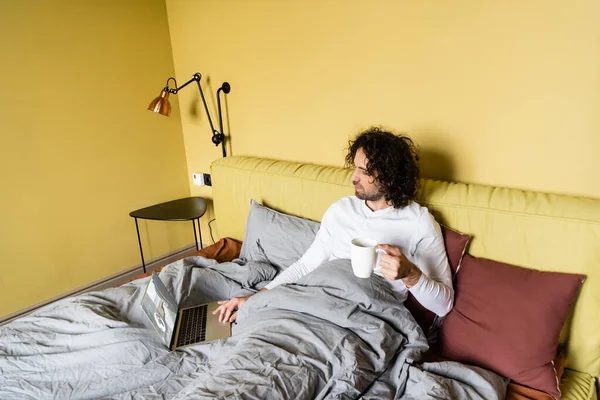 Visão de alto ângulo de jovem freelancer usando laptop com bilhetes site on-line enquanto segurando xícara de café na cama — Fotografia de Stock