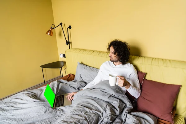 Vista de ángulo alto del joven freelancer utilizando el ordenador portátil con pantalla verde mientras sostiene la taza de café en la cama - foto de stock