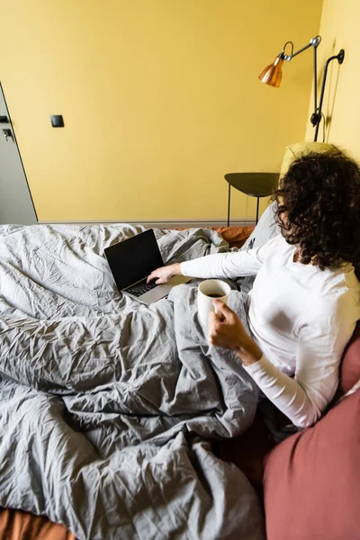 Vista ad alto angolo del freelance riccio utilizzando il computer portatile con schermo bianco mentre si tiene la tazza di caffè a letto — Foto stock