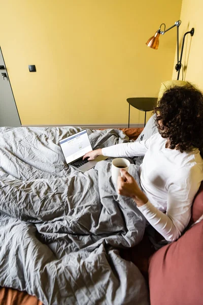 KYIV, UKRAINE - APRIL 25, 2020: high angle view of curly freelancer using laptop with Facebook website while holding cup of coffee in bed — Stock Photo