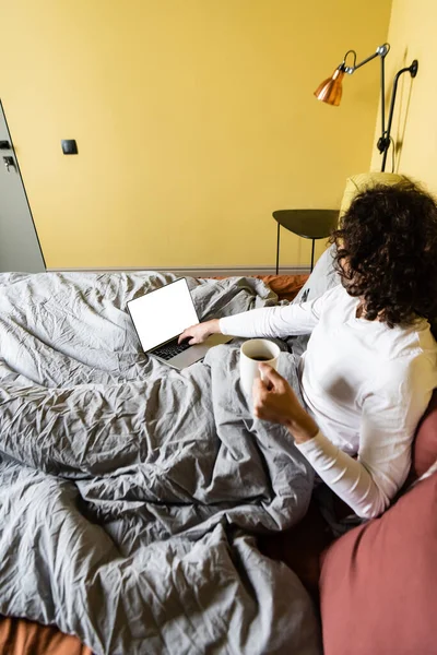 Hochwinkelige Ansicht des lockigen Freelancer mit Laptop mit weißem Bildschirm, während er eine Tasse Kaffee im Bett hält — Stockfoto