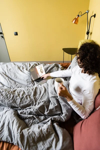 Vista ad alto angolo di riccio freelance in possesso di una tazza di caffè e utilizzando il computer portatile con biglietti sito online a letto — Foto stock