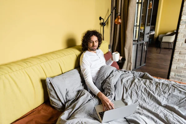 Freelancer encaracolado usando laptop enquanto segurando xícara de café na cama — Fotografia de Stock
