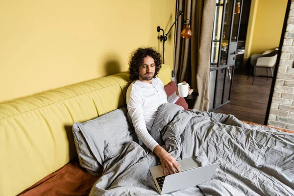 Freelance bouclé à l'aide d'un ordinateur portable tout en tenant une tasse de café au lit — Photo de stock