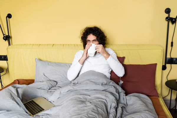 Lockige Freiberuflerin trinkt Kaffee im Bett neben Laptop und blickt in die Kamera — Stockfoto