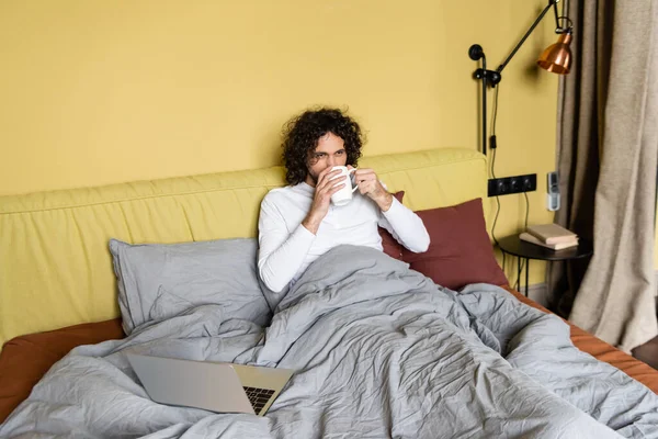 Freelancer encaracolado bebendo café na cama perto do laptop e olhando para longe — Fotografia de Stock