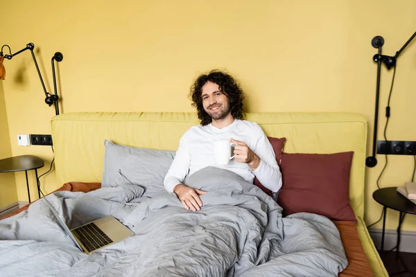 Счастливый фрилансер за чашкой кофе, улыбаясь фотоаппарату в постели рядом с ноутбуком — стоковое фото
