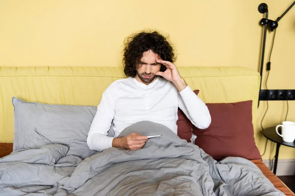 Unzufriedener Mann berührt Kopf beim Blick auf Thermometer im Bett — Stockfoto